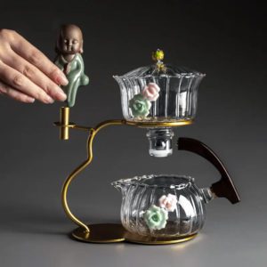 Originale Teekanne aus Glas mit dekorativen Blumendetails und Buddha-Figur auf einem stilvollen goldenen Ständer.