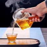 Gehämmerte Glasteekanne mit Holzgriff, die dampfenden Tee in eine kleine Tasse gießt.